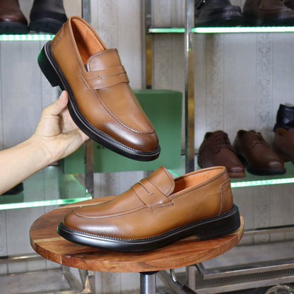 A pair of men's tan leather penny loafers displayed on a wooden stool, with one shoe held up by a hand. The background shows a shoe store interior with glass shelves displaying other footwear.